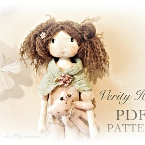 May include: A handmade doll with brown curly hair, wearing a light green shawl and a pink dress, holding a small stuffed animal. The doll is standing in front of a light brown background with the text "Verity Hope PDF PATTERN" in black.