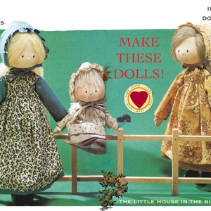 May include: Three handmade dolls with floral print dresses and bonnets stand in front of a wooden fence. The dolls are made with fabric and have painted faces. The text "MAKE THESE DOLLS!" is written in red letters above the dolls. The text "THE LITTLE HOUSE IN THE BIG WOODS" is written below the fence.