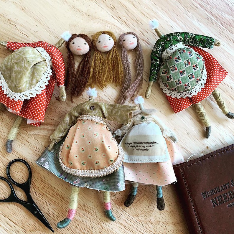 Op de afbeelding: Vijf handgemaakte stoffen poppen met garenhaar. De poppen dragen verschillende gepatroneerde jurken en hebben geborduurde details. De poppen zitten op een houten oppervlak. Een citaat van Leo Buscaglia is gedrukt op een van de jurken van de poppen: "Een enkele roos kan mijn tuin zijn... een enkele vriend mijn wereld."