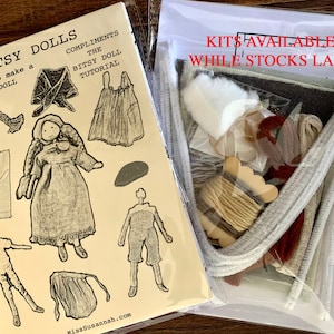 Op de afbeelding: Een kit om een kleine pop te maken, met instructies en materialen. De kit bevat garen, stof, vilt en een houten poppenhoofd. De kit is verpakt in een doorzichtige plastic zak met een witte kaart waarop staat "Kits beschikbaar zolang de voorraad strekt".