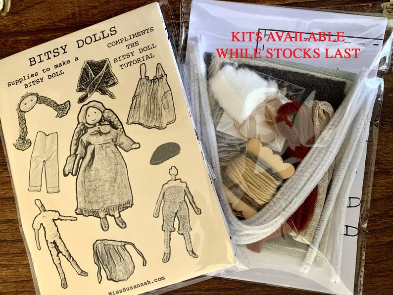 Op de afbeelding: Een kit om een kleine pop te maken, met instructies en materialen. De kit bevat garen, stof, vilt en een houten poppenhoofd. De kit is verpakt in een doorzichtige plastic zak met een witte kaart waarop staat "Kits beschikbaar zolang de voorraad strekt".