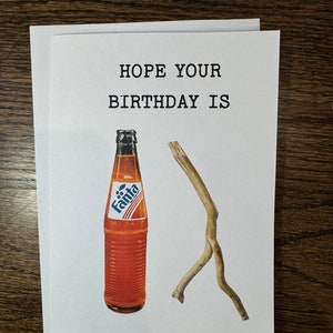 May include: A white card with the text "HOPE YOUR BIRTHDAY IS" printed in black. A bottle of orange soda with the brand name "Fanta" printed on the label is next to a brown twig.