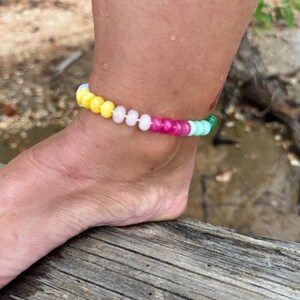 May include: A colorful beaded anklet with yellow, pink, green, and white beads. The anklet is worn on an ankle, with the foot resting on a wooden surface. The beads are round and evenly spaced, creating a vibrant accessory.