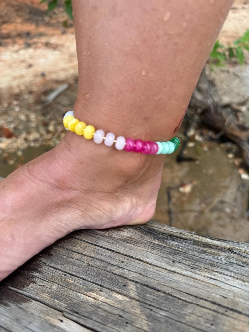 May include: A colorful beaded anklet with yellow, pink, green, and white beads. The anklet is worn on an ankle, with the foot resting on a wooden surface. The beads are round and evenly spaced, creating a vibrant accessory.