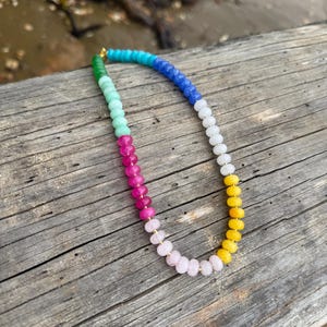May include: A colorful beaded necklace with a gold clasp. The necklace features a gradient of colors, including green, blue, pink, yellow, and white beads. The necklace is displayed on a weathered wooden surface.