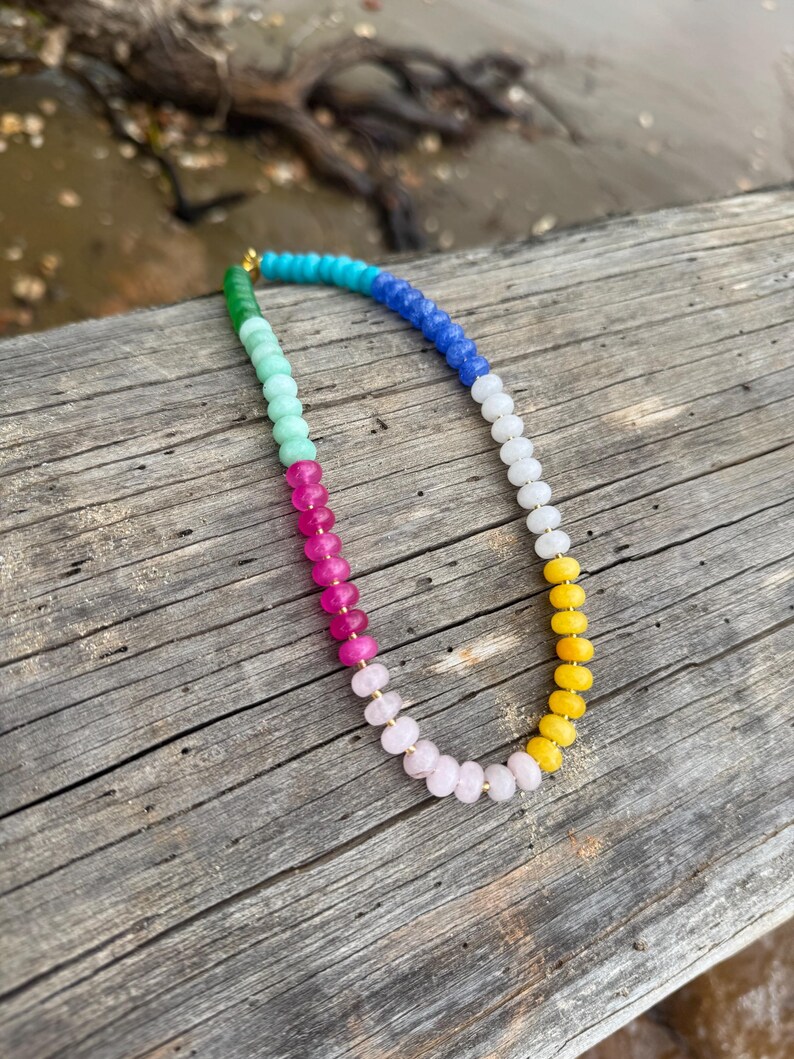 May include: A colorful beaded necklace with a gold clasp. The necklace features a gradient of colors, including green, blue, pink, yellow, and white beads. The necklace is displayed on a weathered wooden surface.