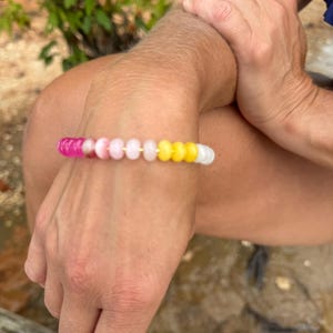 May include: A beaded bracelet with a gradient of colors, including pink, white, and yellow. The bracelet is worn on a wrist, with the beads appearing to be small and round. The background is a natural outdoor setting.