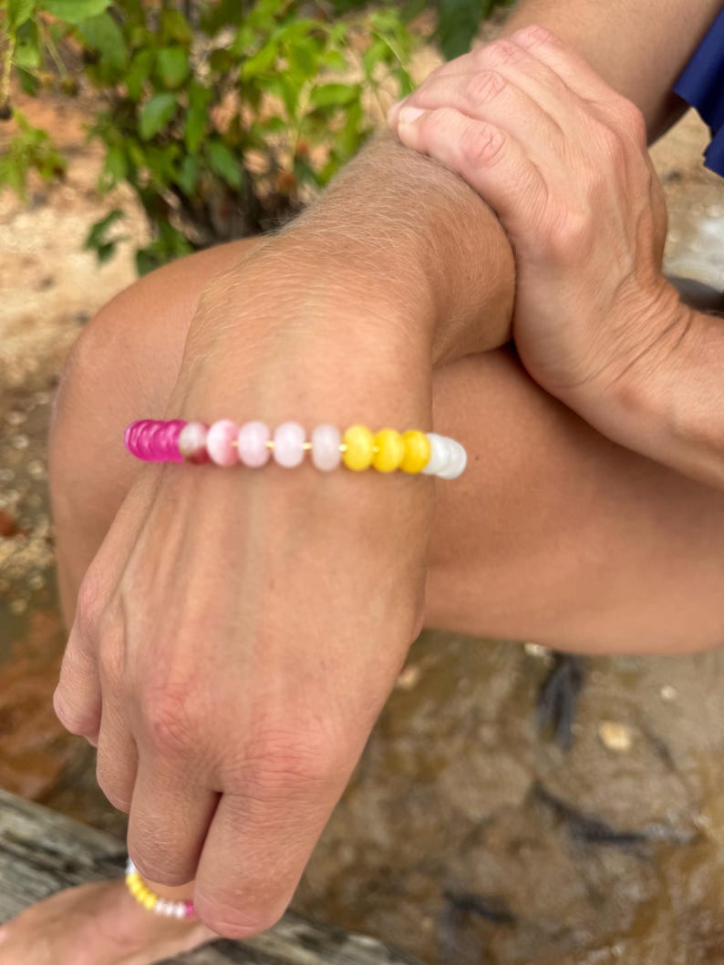 May include: A beaded bracelet with a gradient of colors, including pink, white, and yellow. The bracelet is worn on a wrist, with the beads appearing to be small and round. The background is a natural outdoor setting.