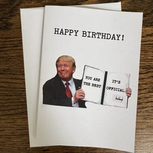 May include: A white birthday card with the text "HAPPY BIRTHDAY!" printed in black. The card features an image of a man holding a sign that reads "YOU ARE THE BEST IT'S OFFICIAL."