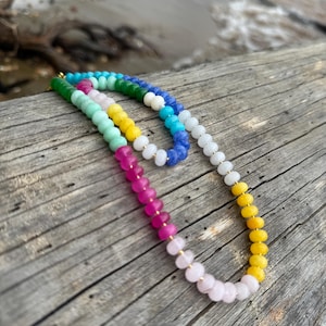 May include: Two beaded necklaces with a gradient of colors, including pink, yellow, green, and blue. The necklaces are resting on a weathered wooden surface, with a blurred background of water and driftwood.