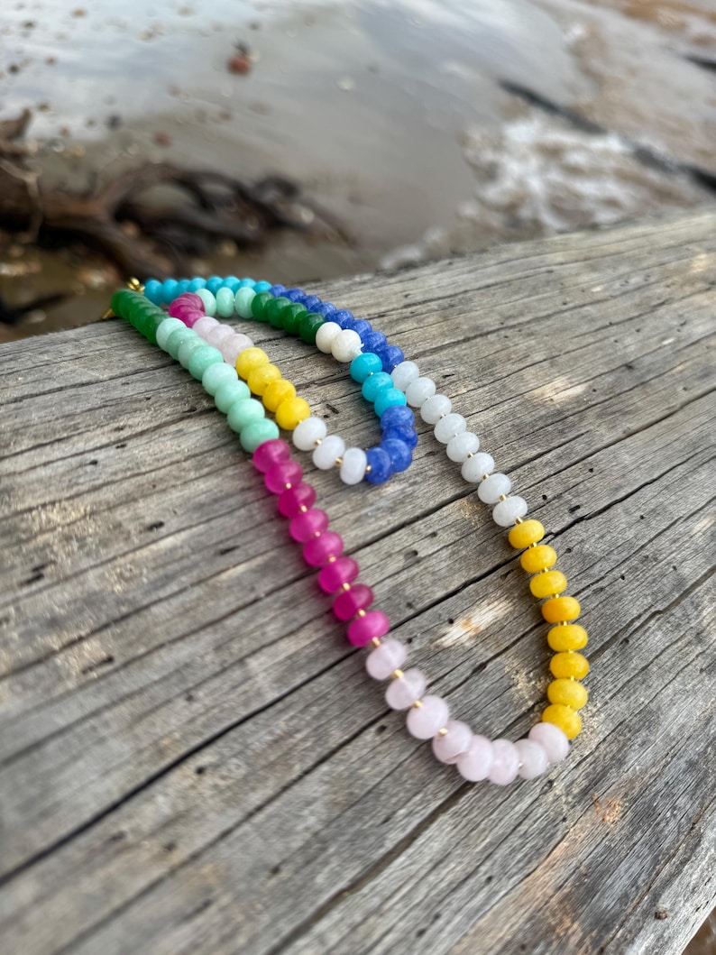 May include: Two beaded necklaces with a gradient of colors, including pink, yellow, green, and blue. The necklaces are resting on a weathered wooden surface, with a blurred background of water and driftwood.
