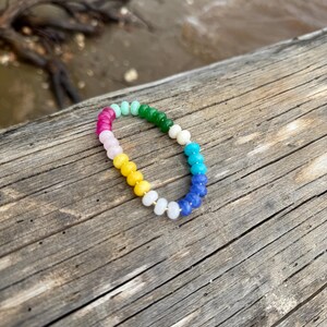 May include: A colorful beaded bracelet with a rainbow gradient of pink, light blue, green, white, yellow, and blue beads. The bracelet is resting on a weathered wooden surface, with a blurred background of water and driftwood.