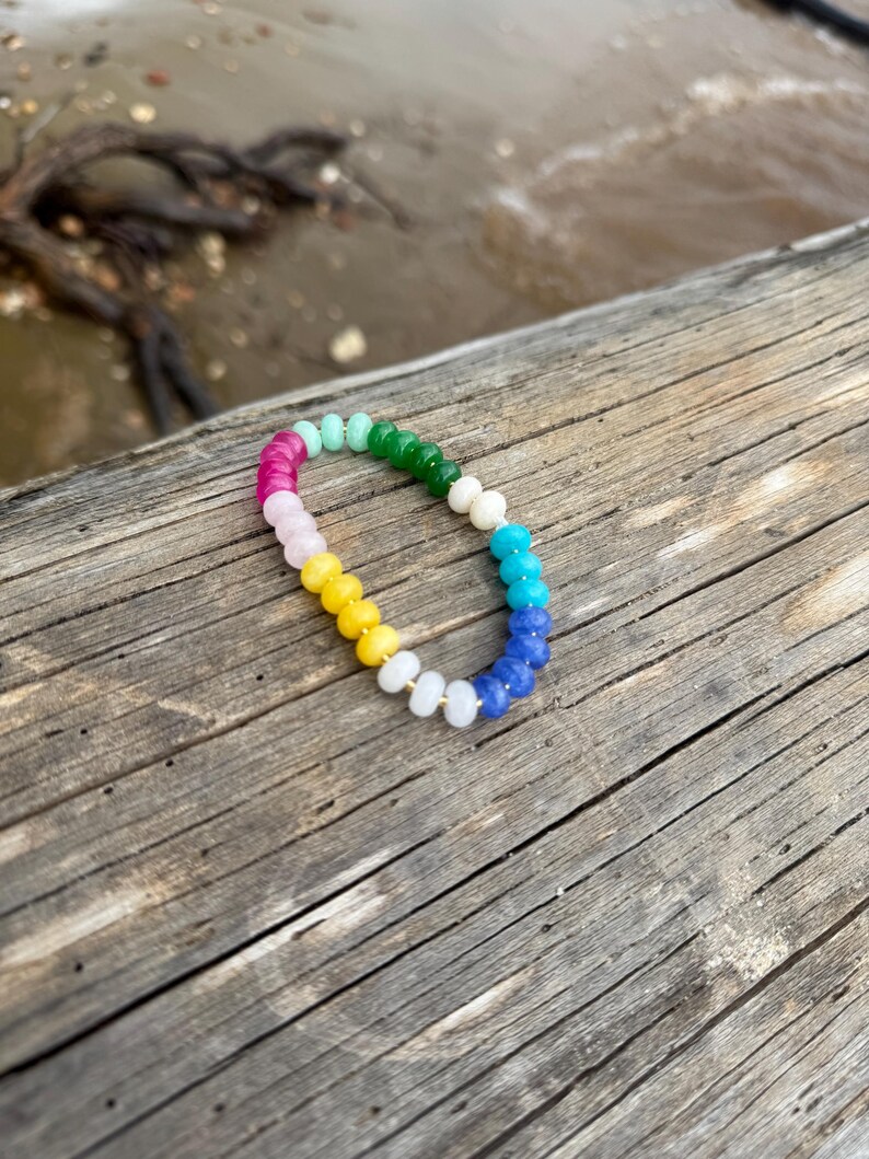 May include: A colorful beaded bracelet with a rainbow gradient of pink, light blue, green, white, yellow, and blue beads. The bracelet is resting on a weathered wooden surface, with a blurred background of water and driftwood.