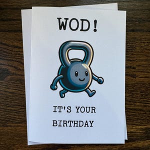 May include: A white card with black text that reads "WOD!" and "IT'S YOUR BIRTHDAY". A cartoon kettlebell with a smiling face is in the center of the card.