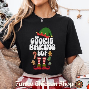 May include: Black t-shirt with the text "The Cookie Baking Elf" in white, featuring elf-themed graphics. The design includes an elf hat, candy cane, gingerbread man, Christmas tree, and striped legs. The shop name "Funky Chicken Shop" is at the bottom.