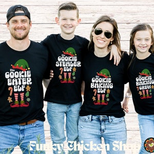 May include: Four black t-shirts with matching designs. Each shirt has a green elf hat, the words "The Cookie Eater Elf", "The Cookie Stealer Elf", "The Cookie Baking Elf", and "The Cookie Baking Crew" with festive graphics.