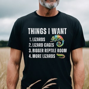 May include: A black t-shirt with white text that reads "THINGS I WANT 1. LIZARDS 2. LIZARD CAGES 3. BIGGER REPTILE ROOM 4. MORE LIZARDS". The shirt features a colorful chameleon and a lizard graphic.