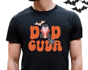 May include: Black t-shirt with the text "DAD CULA" in orange, featuring a vampire graphic and a bat. A spider web graphic is also present. The shirt is a Halloween-themed design.