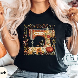 May include: Black t-shirt featuring a colorful illustration of a vintage sewing machine adorned with flowers, leaves, and a pumpkin. The text "Funky Chicken Shop" is at the bottom. The shirt is a classic crew neck.