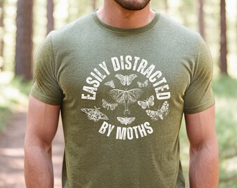May include: Olive green t-shirt with the text "EASILY DISTRACTED BY MOTHS" in a white, circular design. The design includes illustrations of various moths. The shirt is made of a soft material.