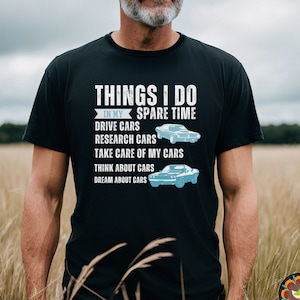 May include: A black t-shirt with white text that reads "Things I do in my spare time: Drive cars, Research cars, Take care of my cars, Think about cars, Dream about cars". The text is surrounded by a blue rectangle. Two blue car silhouettes are shown on the shirt.