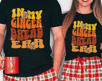 May include: A couple wearing matching black t-shirts with the text "In My Gingerbread Era" in a retro font. The shirts feature a gingerbread man graphic. The couple is wearing red and green plaid pajamas.