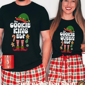 May include: Black t-shirts with elf-themed designs. One shirt says "The Cookie King Elf" and the other says "The Cookie Queen Elf". Both shirts have a green elf hat graphic and gingerbread man, candy cane, and Christmas tree graphics. The models are wearing red plaid pajama pants.