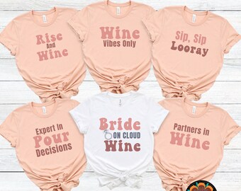 May include: Five peach-colored t-shirts with different text in a retro font. The text on the shirts reads: "Rise and Wine", "Wine Vibes Only", "Sip, Sip Looray", "Expert in Pour Decisions", and "Partners in Wine". One white t-shirt reads "Bride on Cloud Wine".