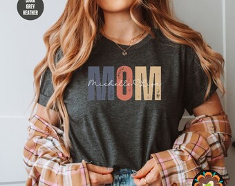 May include: A dark heather grey t-shirt with a vintage-style "MOM" graphic in blue, orange, and yellow. The text "Michelle Lisa" is written in white script below the graphic.