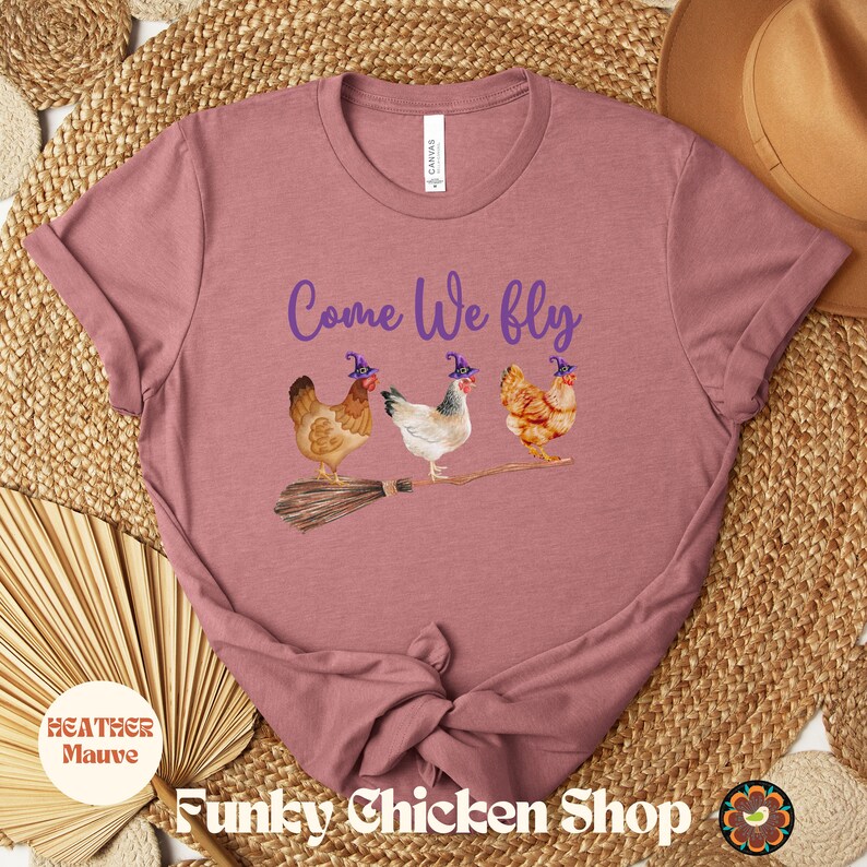 May include: A heather mauve t-shirt with the text "Come We fly" in purple lettering. Three chickens wearing witch hats are flying on a broomstick.