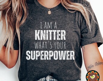 May include: Dark gray t-shirt with the text "I AM A KNITTER WHAT'S YOUR SUPERPOWER" in white. The shirt has a V-neck and is made of a soft material. The shirt is a comfortable and casual wear.