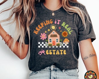 May include: A dark gray t-shirt with a graphic design of a pink house with a checkered pattern, flowers, a mushroom, a sun, and the text "Keeping it Real Estate".