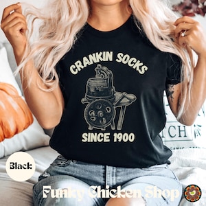May include: Black t-shirt with the text "CRANKIN SOCKS" above an illustration of a vintage sock knitting machine. The text "SINCE 1900" is below the illustration. The words "Funky Chicken Shop" are at the bottom.