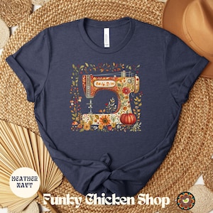 May include: Heather navy blue t-shirt featuring a colorful illustration of a vintage sewing machine adorned with flowers and a pumpkin. The shirt has the text "Funky Chicken Shop" at the bottom.