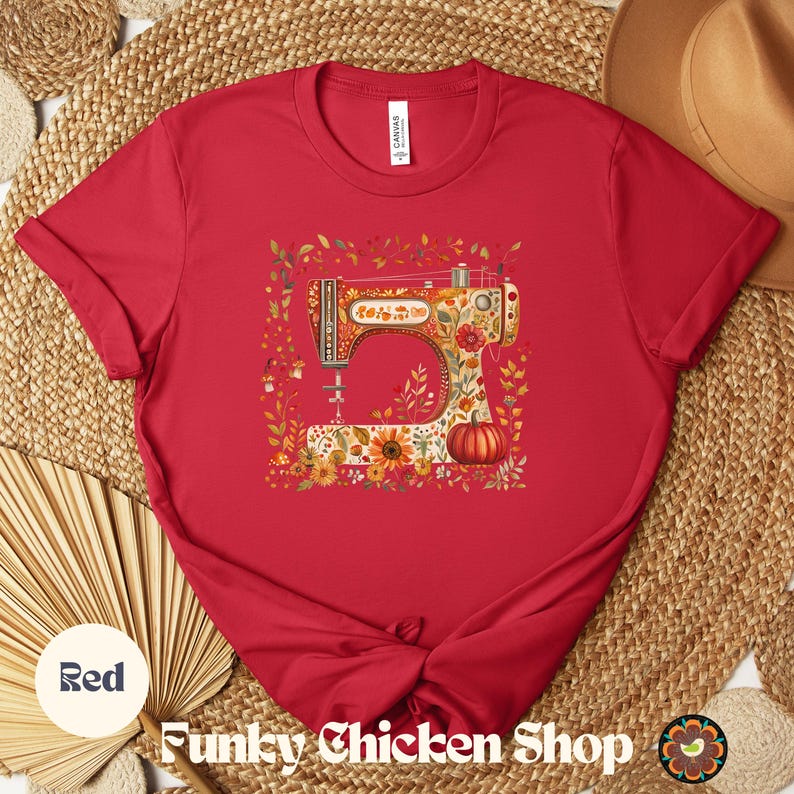 May include: A red t-shirt featuring a colorful illustration of a vintage sewing machine adorned with floral and pumpkin motifs. The shirt has short sleeves and a crew neck. The text "Funky Chicken Shop" is at the bottom.