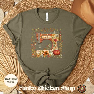 May include: Heather olive t-shirt featuring a colorful illustration of a vintage sewing machine adorned with flowers and a pumpkin. The shirt has the text "Funky Chicken Shop" at the bottom.