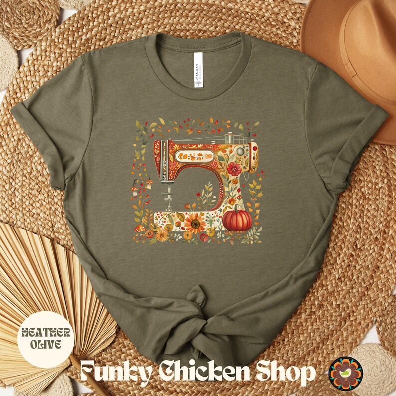 May include: Heather olive t-shirt featuring a colorful illustration of a vintage sewing machine adorned with flowers and a pumpkin. The shirt has the text "Funky Chicken Shop" at the bottom.