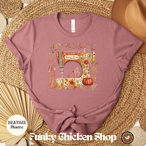 May include: Heather mauve t-shirt featuring a colorful sewing machine design with floral and pumpkin accents. The shirt has the text "Funky Chicken Shop" and "Heather Mauve" printed on it. The design is surrounded by autumn leaves.