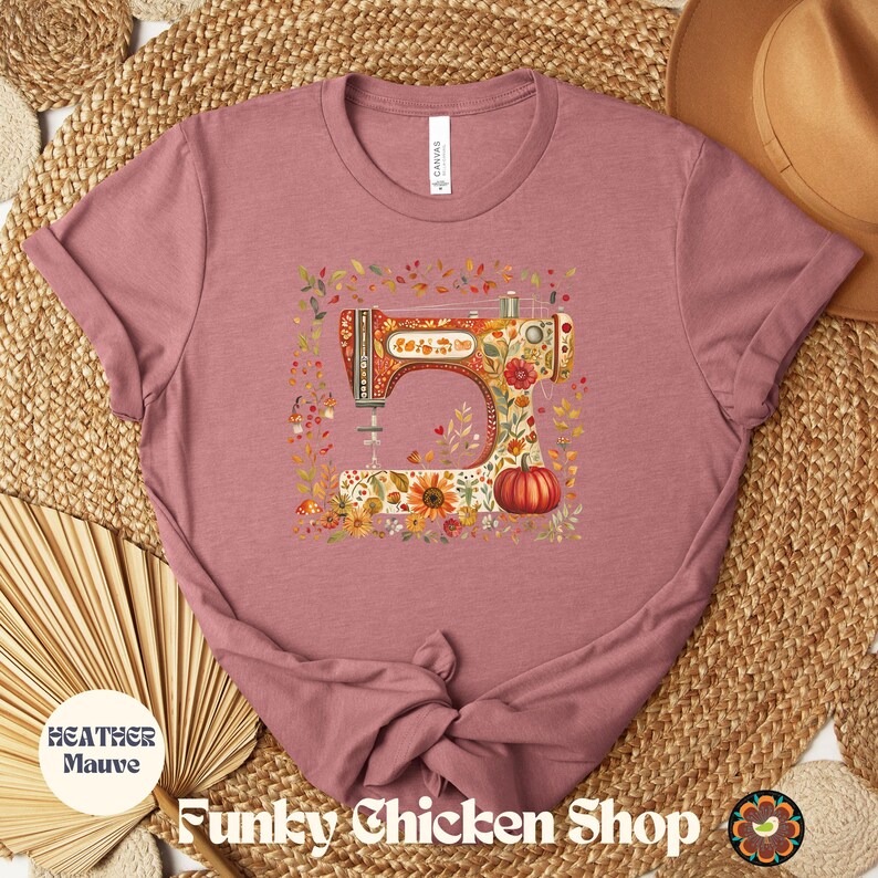 May include: Heather mauve t-shirt featuring a colorful sewing machine design with floral and pumpkin accents. The shirt has the text "Funky Chicken Shop" and "Heather Mauve" printed on it. The design is surrounded by autumn leaves.