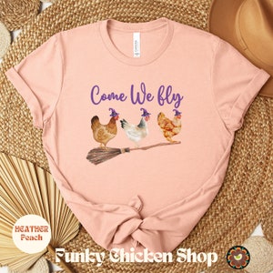 May include: A pink t-shirt with the text "Come We Fly" in purple lettering. Three chickens wearing purple witch hats are riding a broomstick.