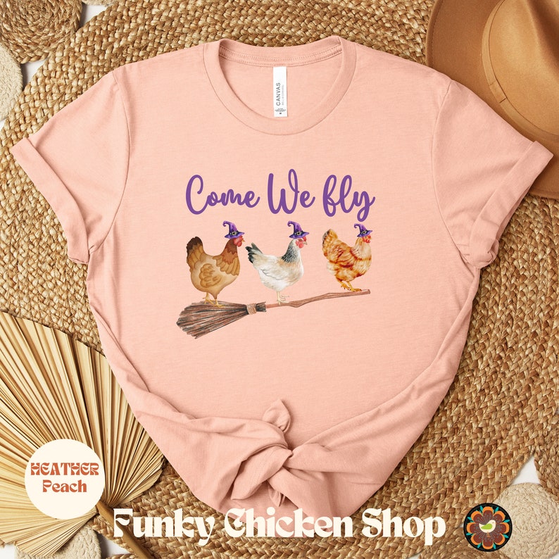 May include: A pink t-shirt with the text "Come We Fly" in purple lettering. Three chickens wearing purple witch hats are riding a broomstick.