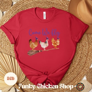 May include: Red t-shirt with the text "Come We Fly" in purple lettering.  Four chickens wearing purple witch hats are flying on a broomstick.