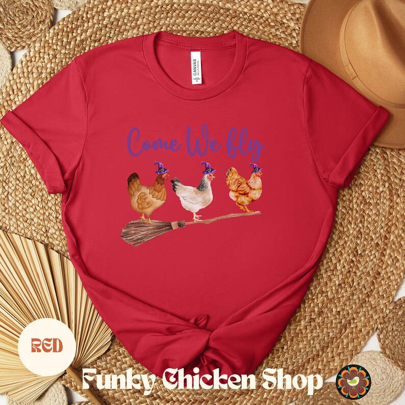 May include: Red t-shirt with the text "Come We Fly" in purple lettering.  Four chickens wearing purple witch hats are flying on a broomstick.