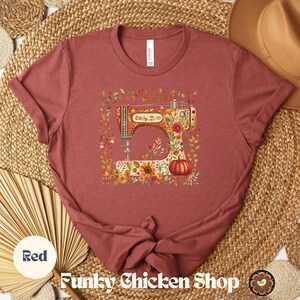 May include: Red t-shirt featuring a colorful illustration of a vintage sewing machine decorated with flowers and a pumpkin. The text "Funky Chicken Shop" is at the bottom. The shirt is made of soft material.
