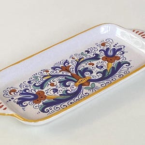 May include: A rectangular ceramic serving tray with rounded corners and two small handles. The tray features a white base with a hand-painted floral pattern in shades of blue, green, and orange, finished with a gold-painted rim and handle edges.