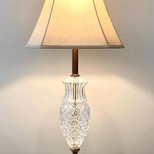 May include: A vintage table lamp with a cream-colored lampshade and a clear, diamond-cut glass base. The lamp has a brass stem and base, and sits on a white marble square. The lamp is designed for indoor use.
