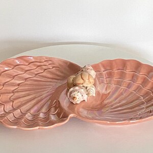May include: A decorative, two-sectioned serving dish in the shape of a seashell. The dish is a soft pink color with iridescent highlights. Several small seashells are clustered in the center. A coastal-themed accent piece.