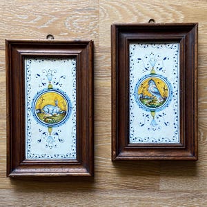 May include: Two framed ceramic tiles with hand-painted designs. One features a rabbit in a yellow circle, the other a bird. Both have blue and yellow accents, set against a white background with a dark brown wooden frame.