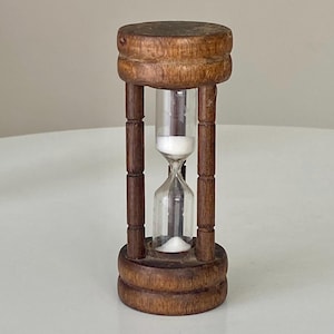 May include: An hourglass featuring a dark brown wooden frame with a cylindrical top and base connected by three vertical pillars. A clear glass bulb is centered within the frame, containing white sand that is currently flowing from top to bottom.