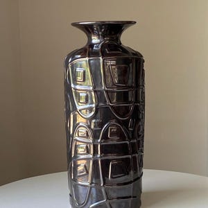 May include: A tall, dark brown ceramic vase with a glossy finish. The vase features a repeating geometric pattern of squares and rectangles. The vase has a flared top and a narrow neck, and it sits on a white surface.